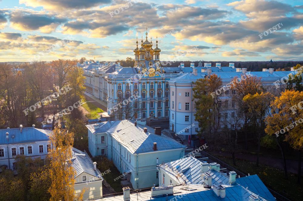 На фотографии изображен Екатерининский дворец в Пушкине, пригороде Санкт-Петербурга. Золотые купола дворца ярко выделяются на фоне осеннего пейзажа и закатного неба. Солнечный свет мягко освещает здание и осенние деревья вокруг. Этот дворец является ярким примером русского барокко и одной из самых известных достопримечательностей России, привлекающей множество туристов со всего мира.